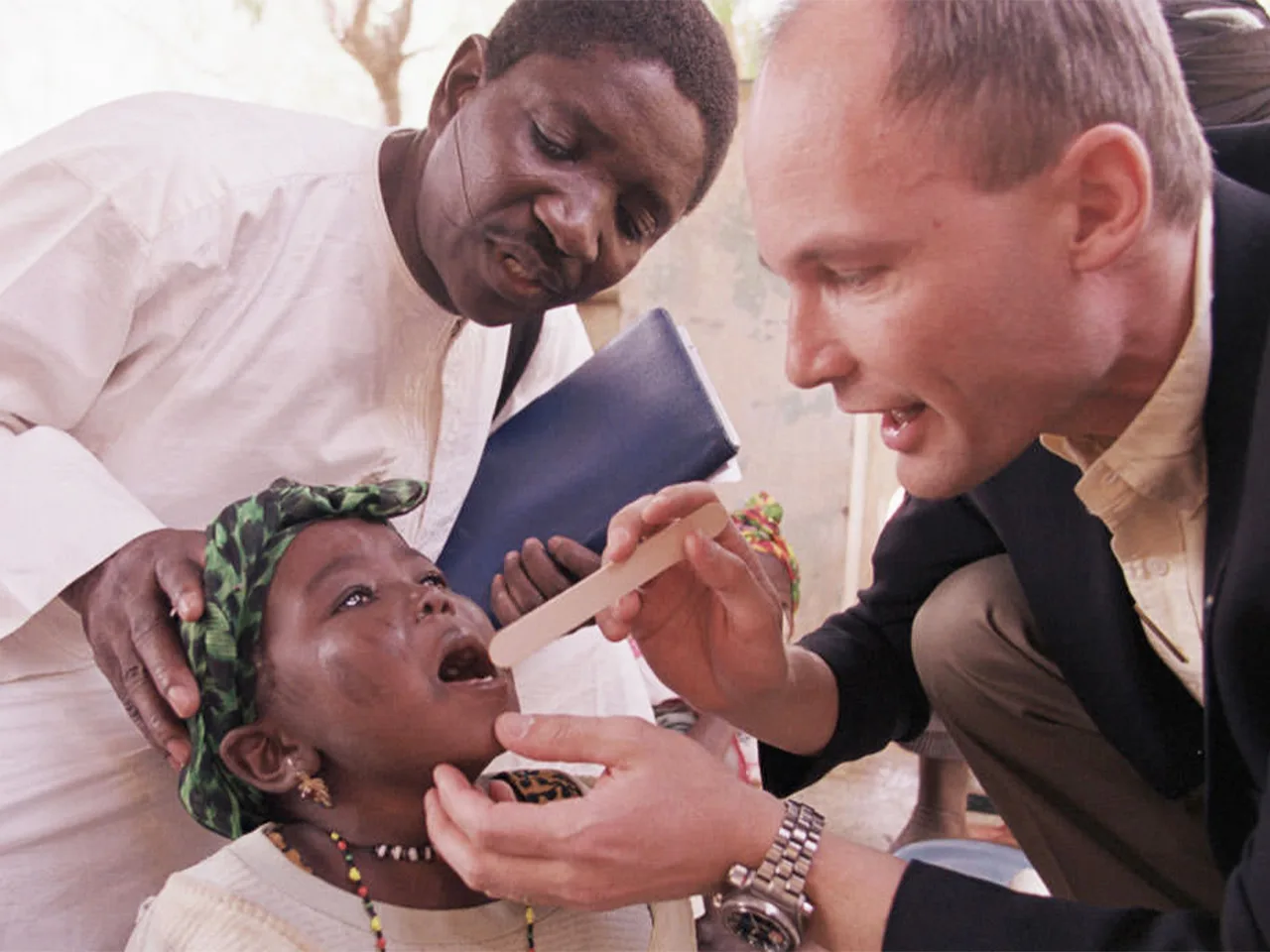 A clinician examines a young girl’s mouth during a noma screening as a health worker supports her.